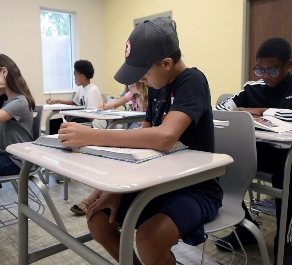 Photo of a boy reading a textbook in a classroom with other students in the background.